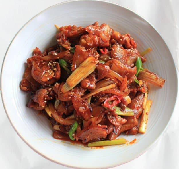 A white bowl of gochujang-marinated pork belly stir-fried with scallions